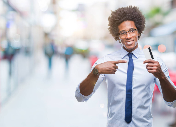 Αφρο αμερικανικό άνθρωπος που κρατά την πιστωτική κάρτα πέρα από το απομονωμένο υπόβαθρο πολύ χαρούμενος, δείχνοντας με το χέρι και το δάχτυλό - Φωτογραφία, εικόνα