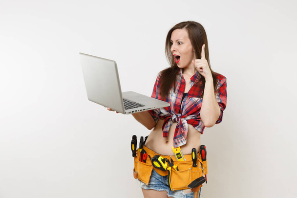 Young pretty handyman woman user in plaid shirt, denim shorts, kit tools belt full of instruments working on laptop pc computer isolated on white background. Female in male work. Renovation concept - 写真・画像
