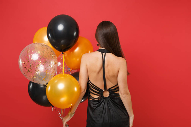 Back rear view of young woman in little black dress celebrating holding air balloons isolated on bright red background. International Women's Day Happy New Year, birthday mockup holiday party concept - Foto, Imagem