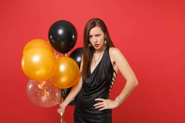 Mujer irritada enojada en vestido negro sosteniendo globos de aire de pie en pose amenazante mientras celebra aislado en el fondo rojo. Día de San Valentín Feliz Año Nuevo, concepto de fiesta de cumpleaños mockup fiesta
 - Foto, imagen