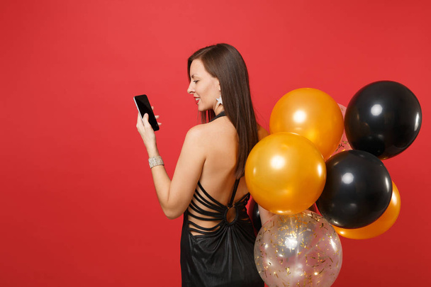 Back rear view of young woman in black dress holding, using mobile phone with blank black empty screen, air balloons isolated on red background. Happy New Year, birthday mockup holiday party concept - Φωτογραφία, εικόνα