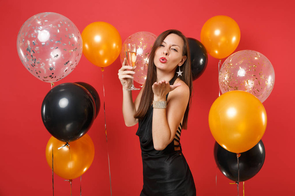 Inschrijving jonge vrouw in zwarte jurk vieren waait lippen, lucht kus verzenden, glas champagne op heldere rode achtergrond lucht ballonnen te houden. Gelukkig Nieuwjaar, concept vakantie partij van verjaardag mockup - Foto, afbeelding