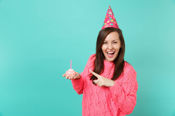 Iloinen nuori nainen neulottu vaaleanpunainen pusero, syntymäpäivä hattu osoittaa etusormella kakku kynttilä kädessä eristetty sininen tausta studio muotokuva. Ihmisten elämäntapa. Mallintaa kopiotilaa
 - Valokuva, kuva