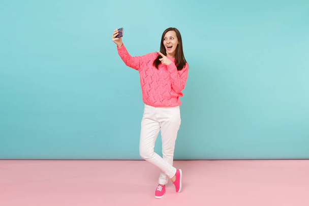 Full length portrait of woman in knitted rose sweater, white pants doing selfie shot on cellphone isolated on bright pink blue pastel wall background. Fashion lifestyle concept. Mock up copy space - Фото, изображение