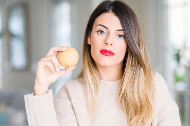 Молода красива жінка тримає свіжу картоплю вдома з впевненим виразом на розумному обличчі думаючи серйозно
 - Фото, зображення