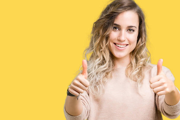 Beautiful young blonde woman wearing sweatershirt over isolated background approving doing positive gesture with hand, thumbs up smiling and happy for success. Looking at the camera, winner gesture. - Photo, Image