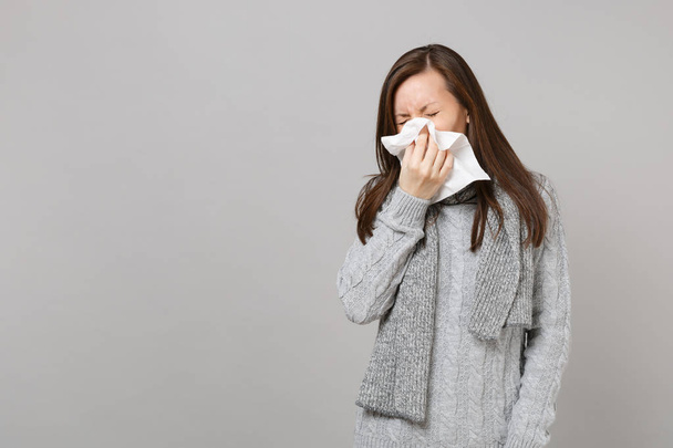 Young woman in gray sweater, scarf having runny nose, blowing nose to napkin isolated on grey wall background. Healthy lifestyle, ill sick disease treatment, cold season concept. Mock up copy space - Photo, image