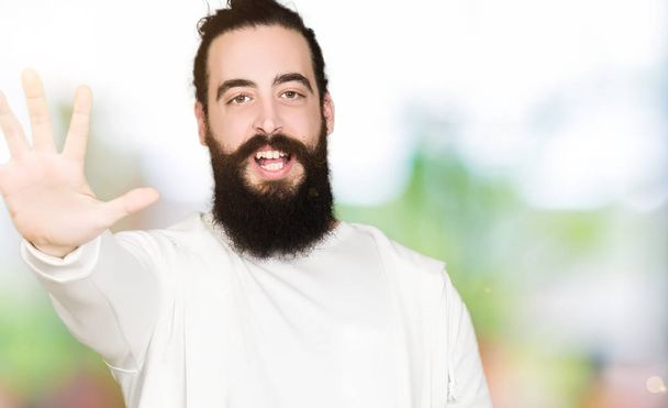 Молодой человек с длинными волосами и бородой в спортивной толстовке показывает и показывает пальцами номер пять улыбаясь уверенно и счастливо
. - Фото, изображение