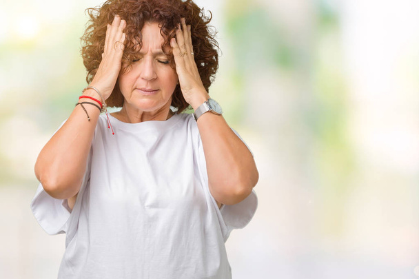 Красивая пожилая женщина среднего возраста в белой футболке на изолированном фоне с рукой на голове от боли в голове из-за стресса. Страдания от мигрени
. - Фото, изображение