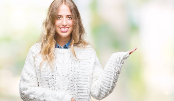 Beautiful young blonde woman wearing winter sweater over isolated background smiling cheerful presenting and pointing with palm of hand looking at the camera. - Photo, Image