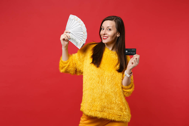 Une jeune femme souriante en pull en fourrure jaune détient une carte bancaire de crédit, fan d'argent en billets de banque en dollars, argent liquide isolé sur fond rouge. Les gens sincères émotions concept de style de vie. Maquette espace de copie
 - Photo, image
