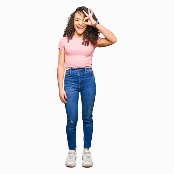Young beautiful woman with curly hair wearing pink t-shirt doing ok gesture with hand smiling, eye looking through fingers with happy face. - Foto, afbeelding