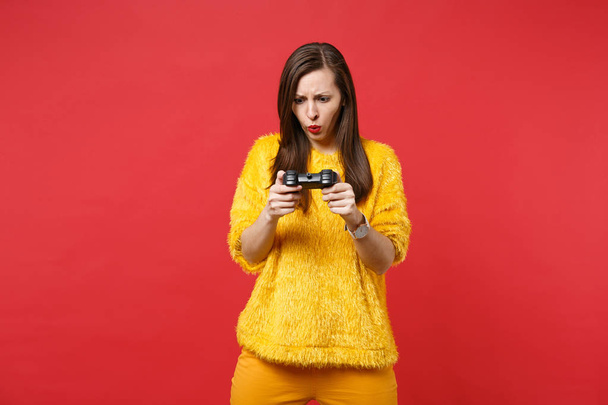 Portrait of puzzled young woman in yellow fur sweater playing video game, looking on joystick isolated on bright red wall background. People sincere emotions, lifestyle concept. Mock up copy space - Фото, зображення