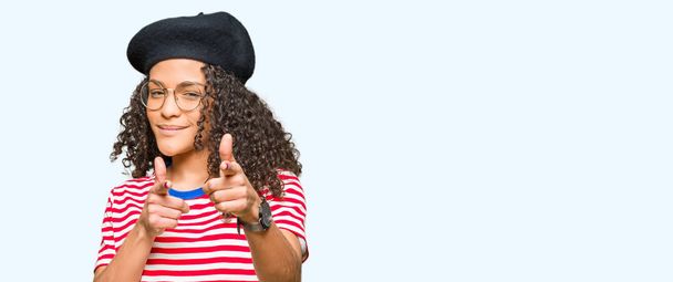 Young beautiful woman with curly hair wearing glasses and fashion beret pointing fingers to camera with happy and funny face. Good energy and vibes. - Photo, Image
