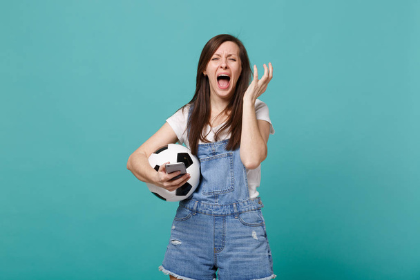 Crying screaming young woman football fan with soccer ball using mobile phone, spreading hands isolated on blue turquoise background. People emotions, sport family leisure concept. Mock up copy space - Photo, Image