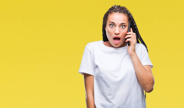 Νέος braided μαλλιών αφρικανικές αμερικανικές κορίτσι καλώντας προβολή χρησιμοποιώντας smartphone πέρα από το απομονωμένο υπόβαθρο φοβάται το σοκ με πρόσωπο έκπληξη, φοβάται και ενθουσιασμένος με φόβο έκφραση - Φωτογραφία, εικόνα
