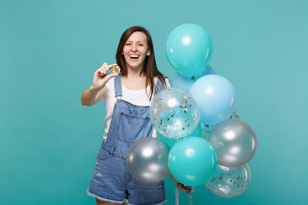 Eine lachende junge Frau in Jeanskleidung hält Bitcoin, die zukünftige Währung, und feiert mit bunten Luftballons auf türkisblauem Hintergrund. Geburtstagsparty, Menschen Emotionen Konzept - Foto, Bild