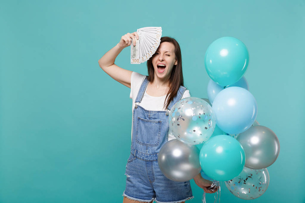Blinkende Frau hält Fan von Geld in Dollarnoten Bargeld in der Nähe Gesicht feiern mit bunten Luftballons isoliert auf blau-türkisfarbenem Hintergrund. Geburtstagsparty, Menschen Emotionen Konzept - Foto, Bild
