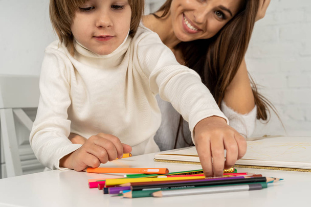 Молодая счастливая мать и маленький сын рисуют цветными карандашами и улыбаются
 - Фото, изображение
