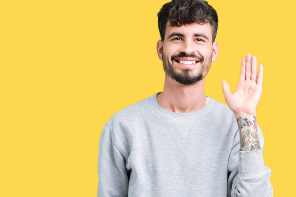 junger gutaussehender Mann, der Sweatshirt über isoliertem Hintergrund trägt und auf ein fröhliches Hallo und eine freundliche Willkommensgeste verzichtet - Foto, Bild