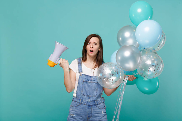 Shocked perplexed young woman keeping mouth open holding megaphone, colorful air balloons while celebrating isolated on blue turquoise wall background. Birthday holiday party, people emotions concept - Photo, Image