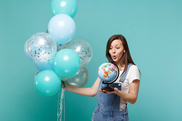 verblüffte junge Frau in Jeanskleidung, die mit bunten Luftballons auf blau-türkisfarbenem Hintergrund auf die Erde blickt. Geburtstagsparty, Menschen Emotionen Konzept - Foto, Bild