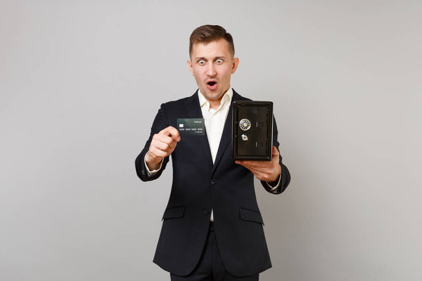 Shocked business man keeping mouth wide open, hold credit bank card, metal bank safe for money accumulation isolated on grey background. Achievement career wealth business concept. Mock up copy space - 写真・画像