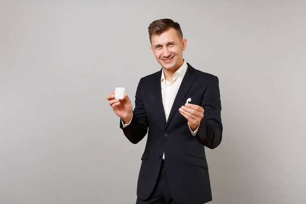Handsome young business man in classic black suit, shirt holding wireless earphones with charging case isolated on grey wall background. Achievement career wealth business concept. Mock up copy space - Photo, Image