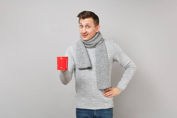 Amazed young man in gray sweater, scarf holding red cup of coffee or tea isolated on grey background in studio. Healthy lifestyle, ill sick disease treatment, cold season concept. Mock up copy space - Photo, Image