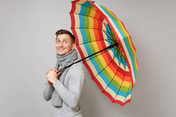 Вид сбоку на молодого человека в сером свитере, шарф держать красочный зонтик глядя вверх изолированы на сером фоне. Здоровый образ жизни людей моды искренние эмоции, концепция холодного сезона. Пространство для копирования
 - Фото, изображение