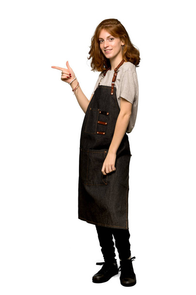 A full-length shot of a Young redhead woman with apron pointing finger to the side in lateral position over isolated white background - Photo, Image