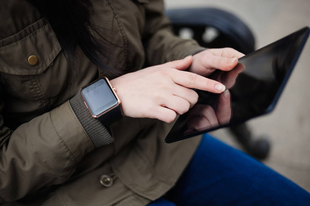 Hipster girl in parka coat using tablet pc with smart watch on her hand. This new gadgets lets you always stay connected to internet and social media networks from anywhere you want. - Фото, изображение
