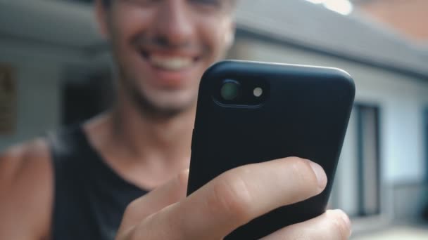 Close up van glimlachen Young Man via Smartphone, browsen in Internet of controleren van sociale netwerken op de binnenplaats of tuin achtergrond - Video