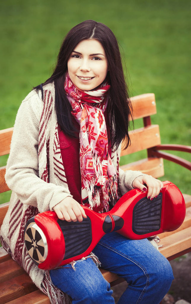 Young white brunette girl holding modern red electric mini  hover board scooter in hands while sitting on a bench in a park. Popular new electric city transport. Girl is wearing trending boho style clothes. - Foto, Imagen
