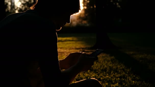 Silhouet van een man met een telefoon in zijn handen bij zonsondergang, de man is het typen van een bericht in een pack op het gras - Video