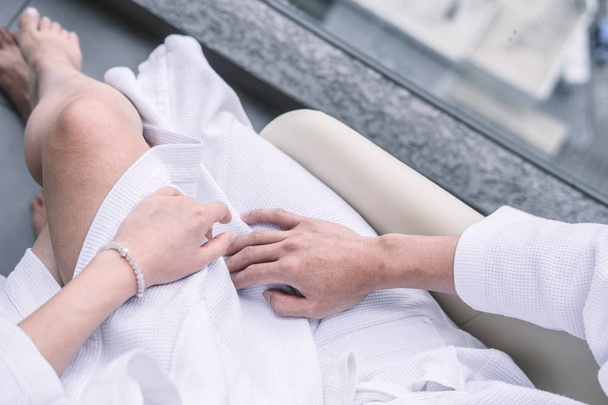Woman sitting on man's laps in white hotel robes touching each other hands. Overhead, man's and woman's legs from above. Intimacy, Valentine's day concept.  - Photo, Image