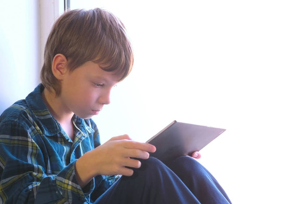 Happy boy is playing games in tablet sitting in windowsill. - Valokuva, kuva