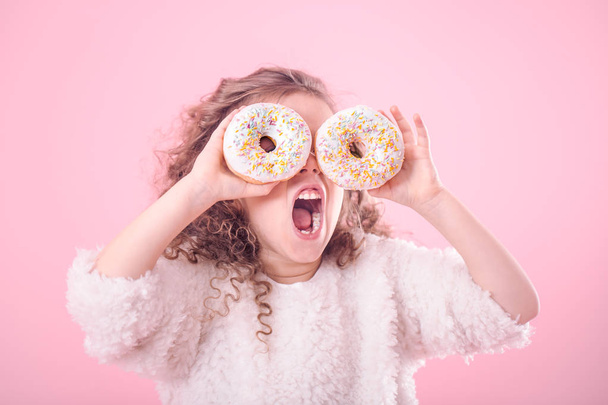 Portrait of a little curly girl with an open mouth and donuts in her hands, closes her eyes with donuts, on a pink background, a place for text - Photo, Image
