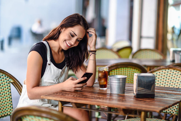 Eine lächelnde junge Frau mit blauen Augen sitzt in einem städtischen Café und benutzt ihr Smartphone. Mädchen mit brauner Wellenfrisur im weißen Jeanskleid. Lifestylekonzept. - Foto, Bild