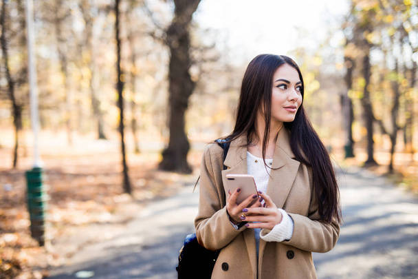 Beautiful woman dressed in autumn coat outdoors, drinking coffee, using mobile phone - Φωτογραφία, εικόνα