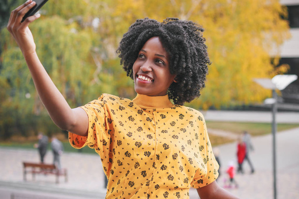 Jeune femme souriante à la peau foncée vêtue de vêtements décontractés faire une photo par son téléphone, fond flou
 - Photo, image