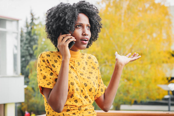 Молодая улыбающаяся темнокожая женщина, одетая в повседневную одежду, разговаривает по телефону, размытый фон
 - Фото, изображение