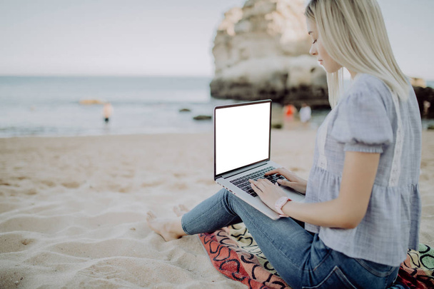 Mujer freelancer trabajando detrás de la computadora portátil sentado en la arena en el fondo de la playa de arena mar, cielo azul y paisaje paradisíaco
. - Foto, imagen