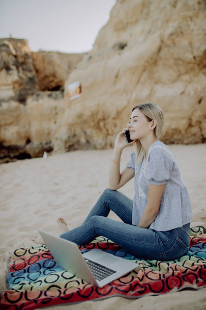 ラップトップが付いている砂の上に座って携帯電話で話しているビーチで働く生産的な若い女性 - 写真・画像