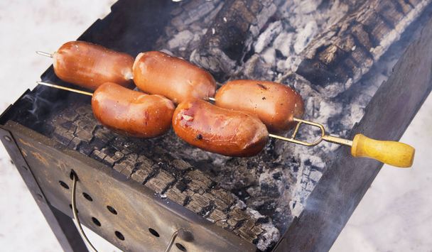 juicy, tasty and fragrant sausages pierced with a skewer with a yellow handle on the grill to roast over the coals at a picnic in the winter forest. Smoke and aroma around. - Photo, Image