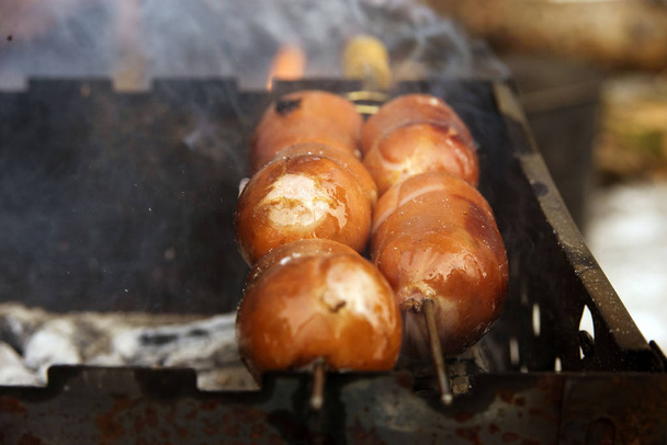 juicy, tasty and fragrant sausages pierced with a skewer with a yellow handle on the grill to roast over the coals at a picnic in the winter forest. Smoke and aroma around. - Photo, Image