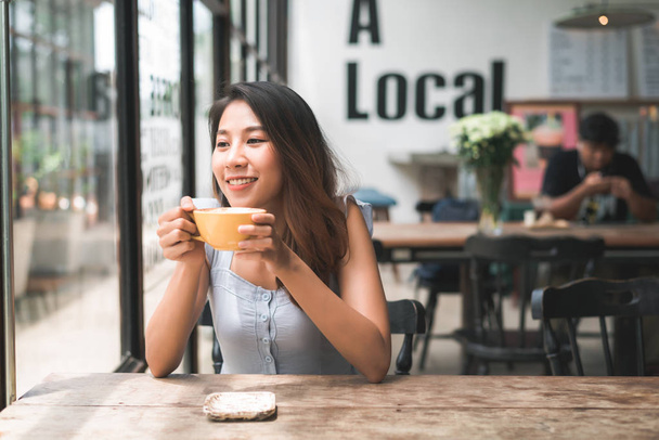 Весела азіатська молода жінка п'є теплу каву або чай, насолоджуючись нею, сидячи в кафе. Приваблива щаслива азіатська жінка тримає чашку кави
. - Фото, зображення