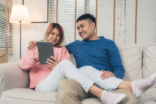 Attractive Asian sweet couple using tablet while lying on the sofa when relax in their living room at home. Husband and his wife using relax and romantic time at home concept. - Valokuva, kuva