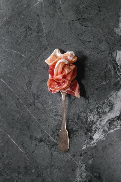 Antipasto meat assorti of sliced jamon, salami, chorizo sausage on fork over black marble background. Flat lay, space. Appetizer concept - Photo, image