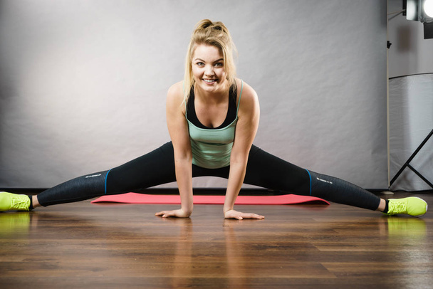 Young blonde woman in sportswear sitting on wooden floor indoor stretching legs doing twine. Training at home, being fit and healthy. - Foto, afbeelding